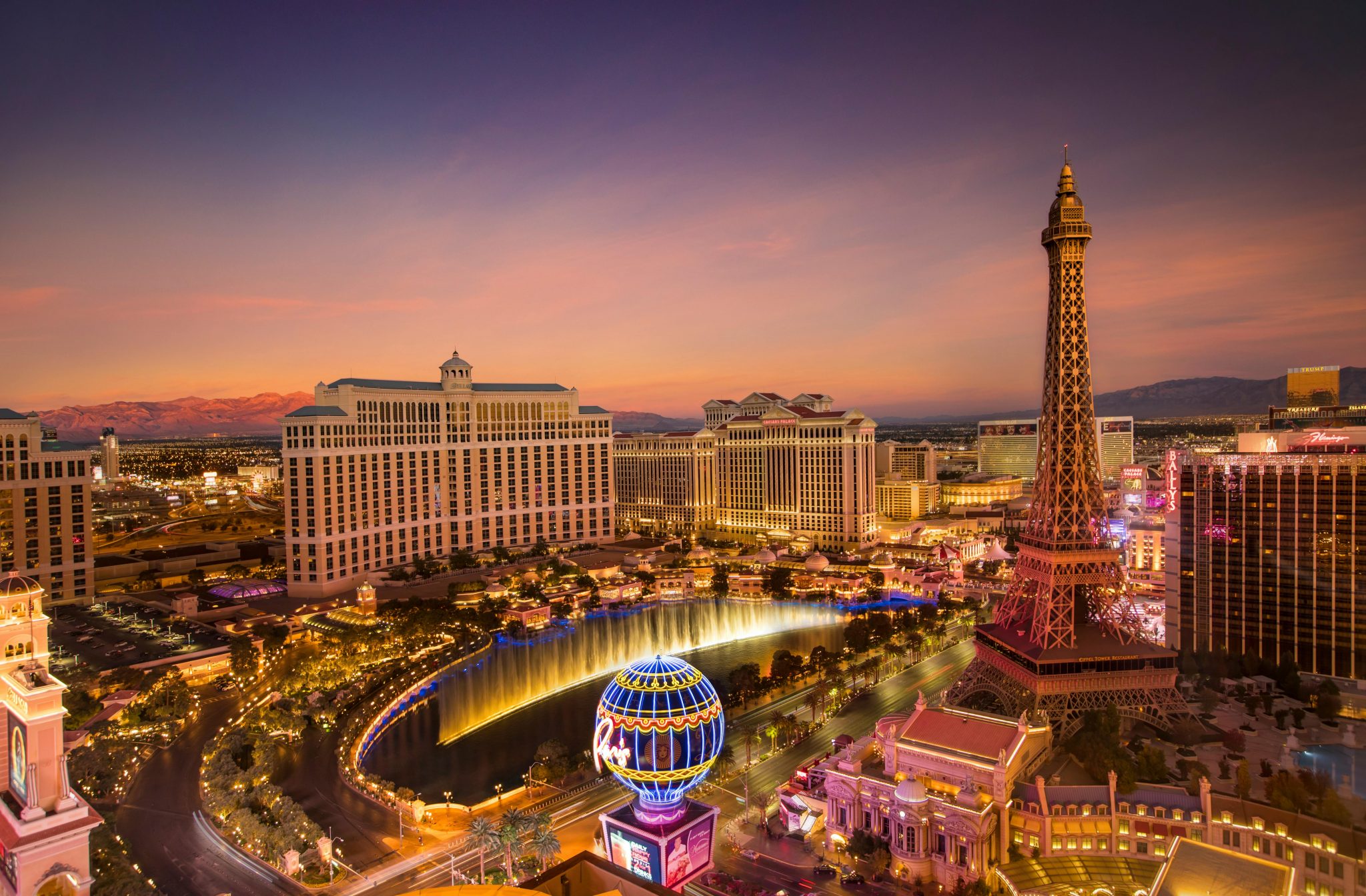 A vibrant night view of the Las Vegas Strip showcasing iconic landmarks like the Bellagio fountains, Paris Las Vegas Eiffel Tower replica, and the Stratosphere Casino. The scene is lit by neon lights, with busy streets filled with cars and tourists, capturing the excitement and glamour of Las Vegas nightlife.